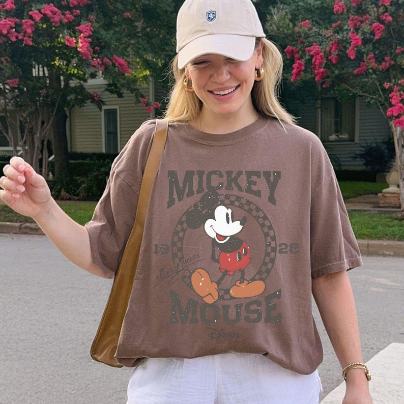 Puede incluir: Camiseta marr&oacute;n con un gr&aacute;fico desgastado de Mickey Mouse. El dise&ntilde;o incluye las palabras "Mickey Mouse" y los a&ntilde;os "1928" y "Disney". La camiseta tiene mangas cortas y un corte holgado.