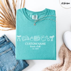 May include: A seafoam green t-shirt featuring a white graphic of bows, an open book, a coffee mug, a stack of books, a closed book, and glasses. The text reads CUSTOM NAME book club est. 2026. A silver chain necklace rests on the neckline.