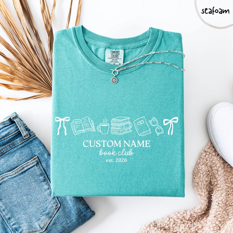 May include: A seafoam green t-shirt featuring a white graphic of bows, an open book, a coffee mug, a stack of books, a closed book, and glasses. The text reads CUSTOM NAME book club est. 2026. A silver chain necklace rests on the neckline.