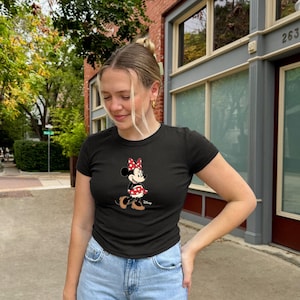 May include: Black crew neck t-shirt featuring a Minnie Mouse graphic. The graphic shows Minnie Mouse wearing a red bow with white polka dots and a matching skirt. The shirt is paired with blue jeans.