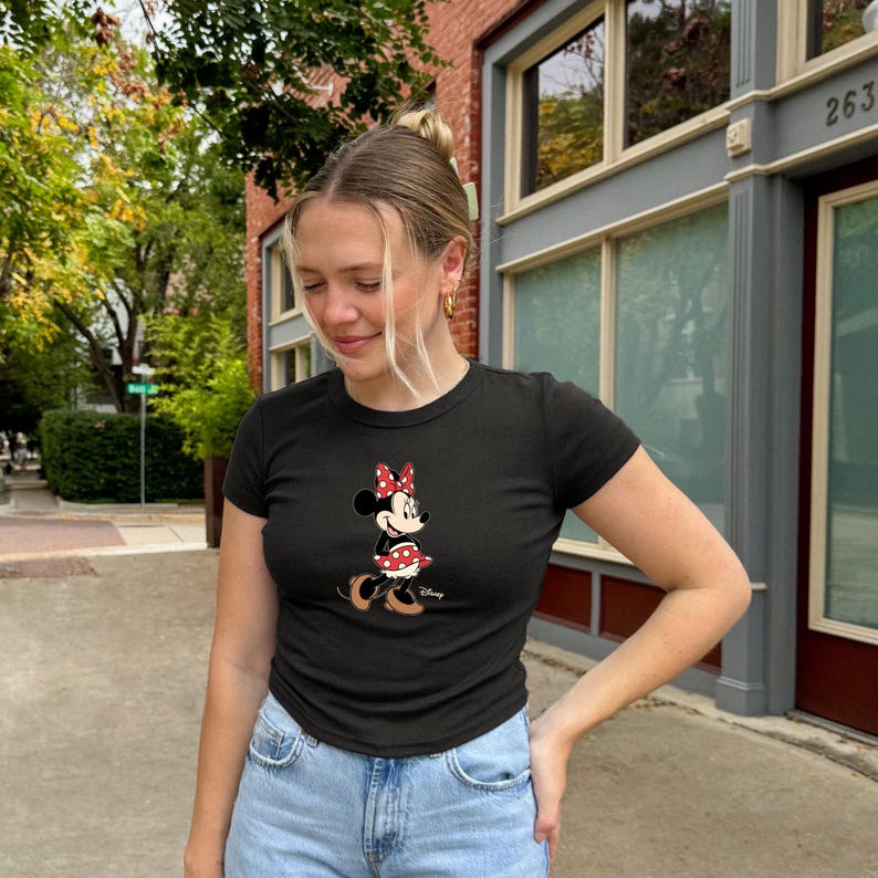 May include: Black crew neck t-shirt featuring a Minnie Mouse graphic. The graphic shows Minnie Mouse wearing a red bow with white polka dots and a matching skirt. The shirt is paired with blue jeans.