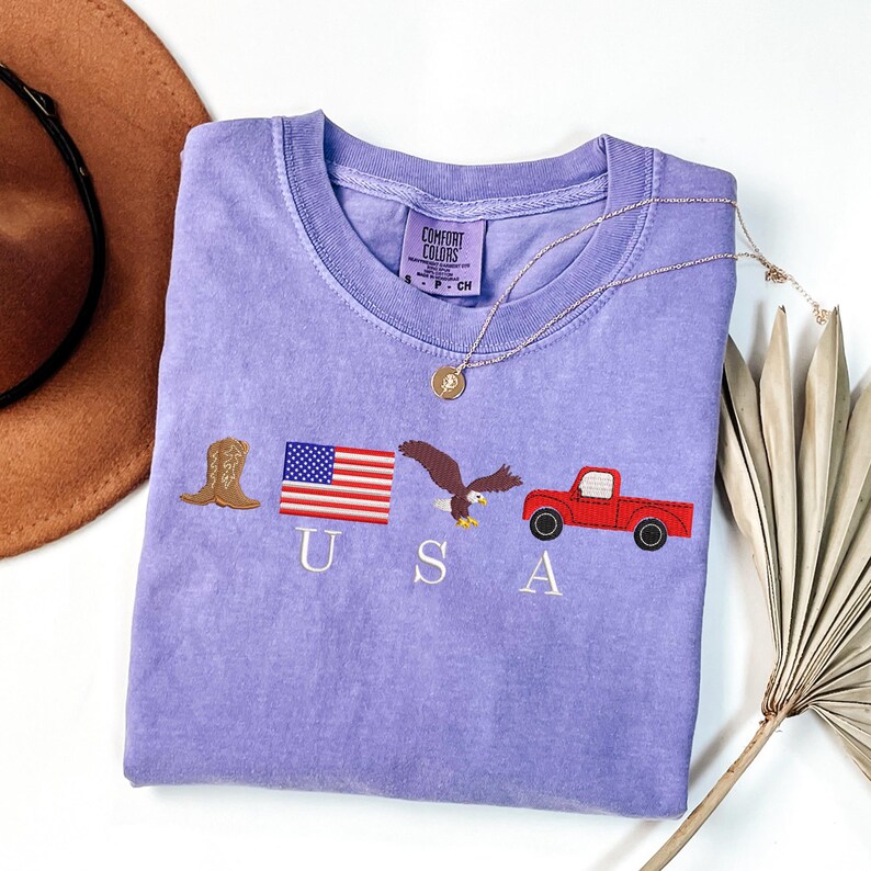 May include: A light purple Comfort Colors t-shirt with embroidered patriotic symbols: a cowboy boot, American flag, eagle, and red truck. The word "USA" is embroidered below the symbols. A gold necklace is draped across the shirt.