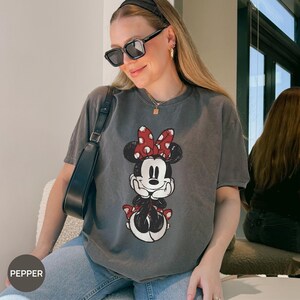 Puede incluir: Una camiseta gris con un gr&aacute;fico de Minnie Mouse con un lazo rojo y blanco de lunares. La ilustraci&oacute;n es de estilo vintage. La camiseta la lleva una persona con gafas de sol y una diadema negra.