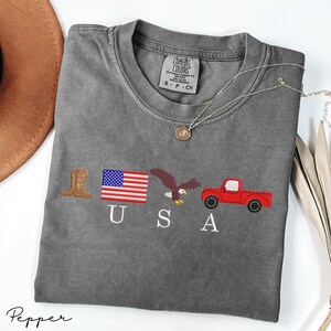 May include: A gray Comfort Colors t-shirt with embroidered patriotic symbols: a cowboy boot, American flag, eagle, and red truck. The word "USA" is embroidered below the symbols. A silver necklace with a pendant is on the shirt.