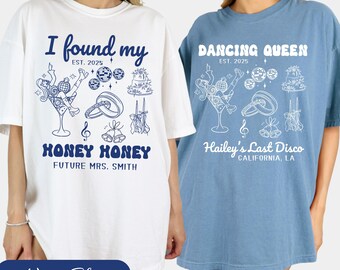 May include: Two white t-shirts with blue text and illustrations. The first shirt says "I found my Honey Honey" with a wedding-themed illustration. The second shirt says "Dancing Queen" with a disco-themed illustration.