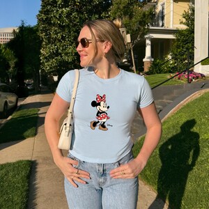 May include: Pale blue t-shirt with a Minnie Mouse design. The cartoon character is wearing a red and white polka dot skirt and bow. The top has short sleeves and a round neckline.