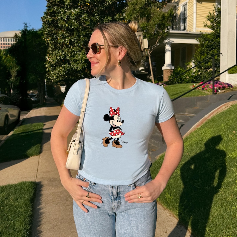 May include: Pale blue t-shirt with a Minnie Mouse design. The cartoon character is wearing a red and white polka dot skirt and bow. The top has short sleeves and a round neckline.