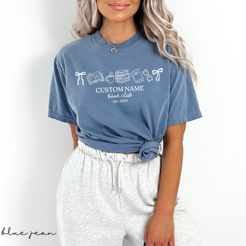 May include: A blue short-sleeve t-shirt featuring a white graphic of bows, books, a coffee mug, and glasses. The text reads CUSTOM NAME book club est. 2026. The shirt is paired with light gray sweatpants with a drawstring waist and side pockets.