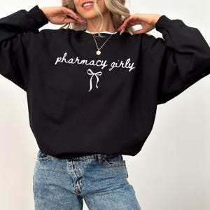 Puede incluir: Sudadera negra con la frase "pharmacy girly" en escritura cursiva y un lazo gr&aacute;fico. La sudadera tiene un cuello blanco y se combina con vaqueros azules.