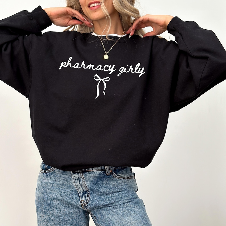 Puede incluir: Sudadera negra con la frase "pharmacy girly" en escritura cursiva y un lazo gr&aacute;fico. La sudadera tiene un cuello blanco y se combina con vaqueros azules.