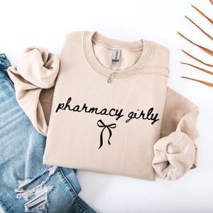 Puede incluir: Sudadera beige de cuello redondo con las palabras "pharmacy girly" en fuente negra y un lazo negro. Un collar plateado est&aacute; sobre la sudadera. La sudadera est&aacute; doblada junto a unos vaqueros azules.