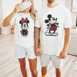 Puede incluir: Dos camisetas blancas a juego con personajes de dibujos animados. Una camiseta tiene un gr&aacute;fico de Minnie Mouse con un lazo y un vestido rojos. La otra camiseta tiene un gr&aacute;fico de Mickey Mouse con pantalones cortos rojos. Ambas camisetas se combinan con pantalones cortos grises.