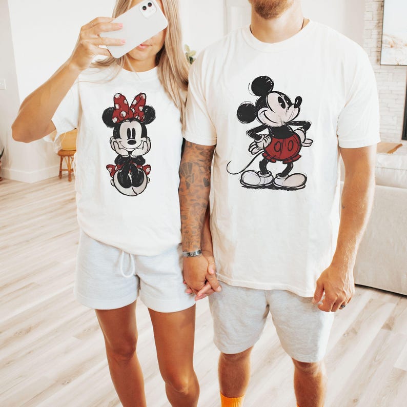 Puede incluir: Dos camisetas blancas a juego con personajes de dibujos animados. Una camiseta tiene un gr&aacute;fico de Minnie Mouse con un lazo y un vestido rojos. La otra camiseta tiene un gr&aacute;fico de Mickey Mouse con pantalones cortos rojos. Ambas camisetas se combinan con pantalones cortos grises.