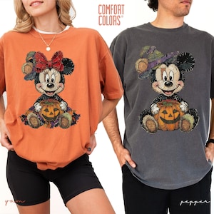 May include: Two short-sleeve crew neck t-shirts, one in orange and one in dark gray, each featuring a graphic of a stitched-style cartoon mouse sitting with a jack-o'-lantern. The orange shirt displays a mouse with a red bow, while the gray shirt shows a mouse wearing a purple witch hat. The text COMFORT COLORS is visible.
