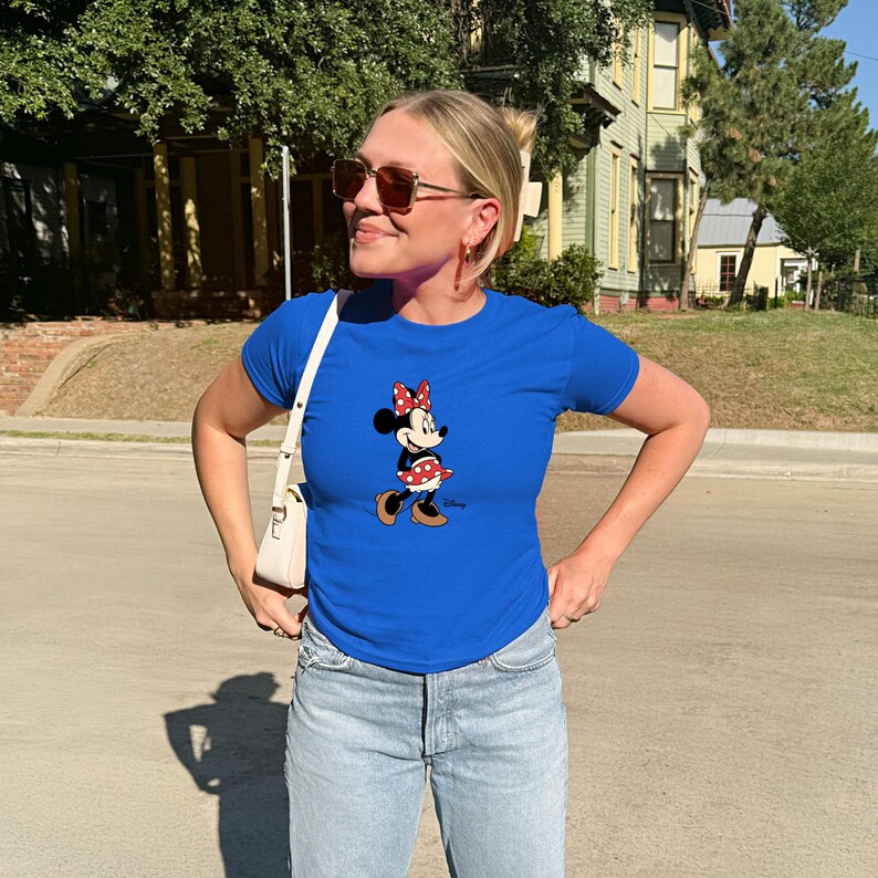 May include: A bright blue t-shirt with a Minnie Mouse design. Minnie is shown with a red bow, a polka dot skirt, and brown shoes. The shirt is worn with light wash jeans and a white handbag.