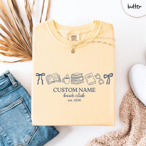 May include: A butter yellow short-sleeve t-shirt featuring a dark blue graphic of two bows, an open book, a coffee mug, a stack of books, a closed book, and glasses. The text reads CUSTOM NAME book club est. 2026 in a dark blue font.