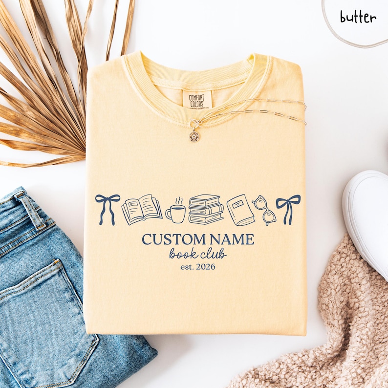 May include: A butter yellow short-sleeve t-shirt featuring a dark blue graphic of two bows, an open book, a coffee mug, a stack of books, a closed book, and glasses. The text reads CUSTOM NAME book club est. 2026 in a dark blue font.