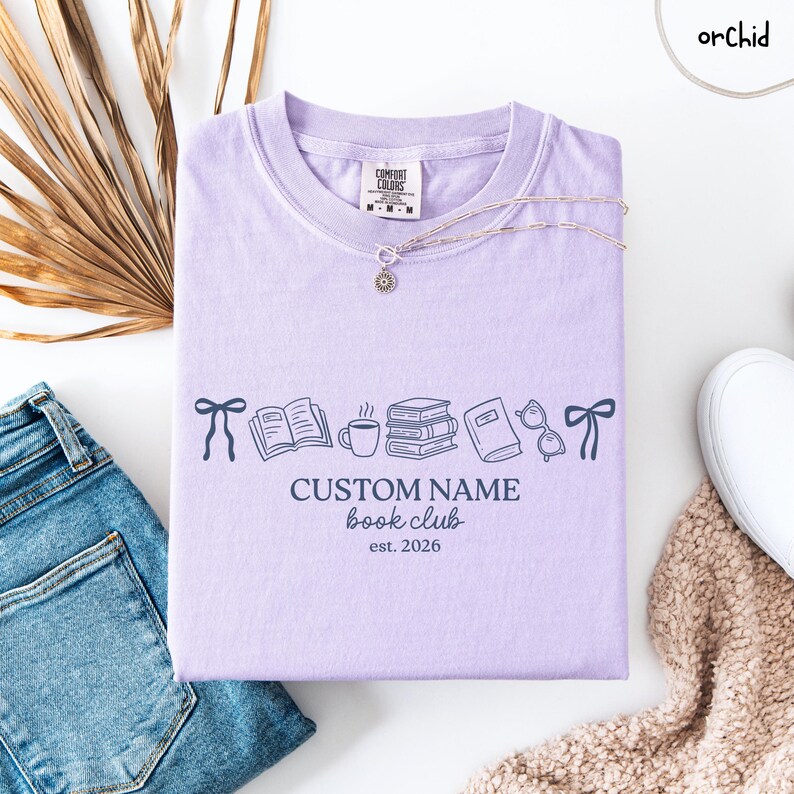 May include: A light purple t-shirt featuring a dark blue graphic print of bows, an open book, a coffee mug, a stack of books, a closed book, and glasses. The text reads CUSTOM NAME book club est. 2026. The shirt has a Comfort Colors brand label.