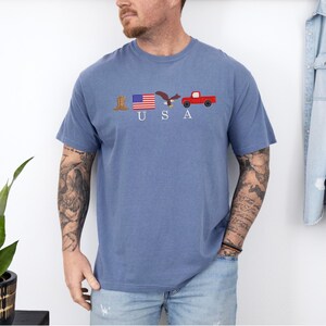 May include: Blue short-sleeved t-shirt with the word "USA" and embroidered patriotic symbols: a cowboy boot, American flag, eagle, and red pickup truck. The shirt is made of a soft, comfortable material.