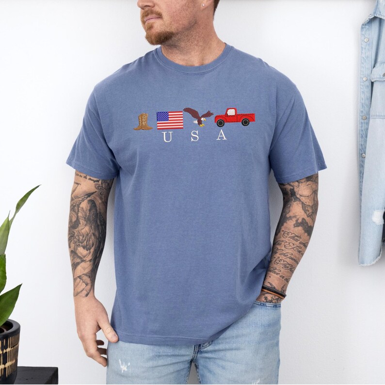 May include: Blue short-sleeved t-shirt with the word "USA" and embroidered patriotic symbols: a cowboy boot, American flag, eagle, and red pickup truck. The shirt is made of a soft, comfortable material.