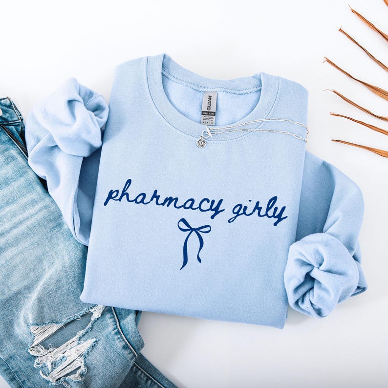 Puede incluir: Sudadera azul claro con las palabras "pharmacy girly" en escritura azul oscuro y un lazo. Un collar plateado con un peque&ntilde;o colgante est&aacute; sobre la sudadera. Se ven unos vaqueros azul claro a la izquierda.