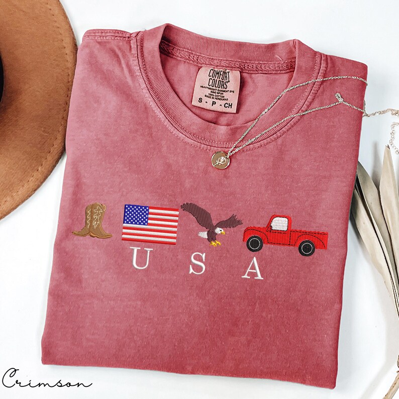 May include: Crimson-colored t-shirt with embroidered USA-themed graphics: a cowboy boot, American flag, eagle, and red truck. The shirt has a crew neck and a small tag. A silver necklace with a pendant is draped across the shirt.