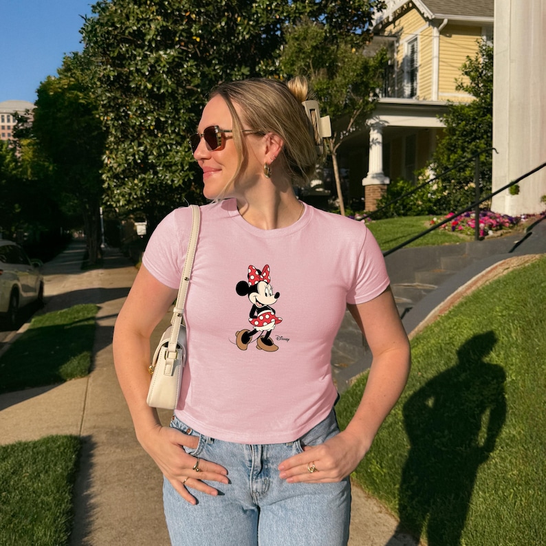 May include: A pale pink t-shirt with a Minnie Mouse design. The cartoon character is shown in black, red, and white, with a red polka-dot skirt and bow. The shirt is worn with blue jeans.