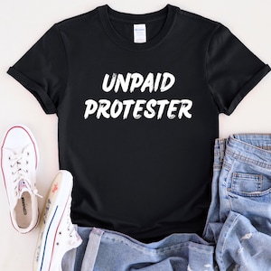May include: Black t-shirt with white text that reads "UNPAID PROTESTER". The shirt is rolled up at the sleeves and is laying on a white surface with a pair of white sneakers and blue jeans.