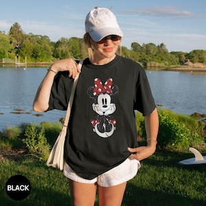 Puede incluir: Camiseta negra con un dise&ntilde;o desgastado de Minnie Mouse con un lazo rojo de lunares. El dise&ntilde;o incluye la cara y la parte inferior del cuerpo de Minnie. La palabra "BLACK" est&aacute; impresa en un c&iacute;rculo negro. La camiseta la lleva una persona.