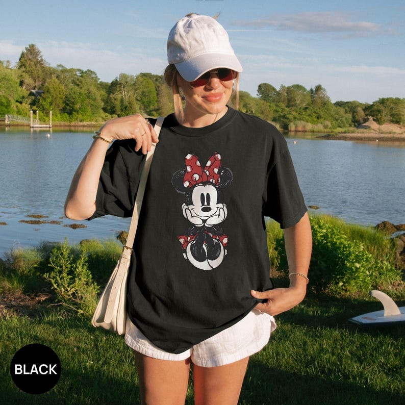 Puede incluir: Camiseta negra con un dise&ntilde;o desgastado de Minnie Mouse con un lazo rojo de lunares. El dise&ntilde;o incluye la cara y la parte inferior del cuerpo de Minnie. La palabra "BLACK" est&aacute; impresa en un c&iacute;rculo negro. La camiseta la lleva una persona.