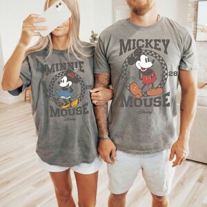 Puede incluir: Camisetas grises con Mickey y Minnie Mouse. La camiseta de Mickey Mouse tiene un gr&aacute;fico de Mickey con pantalones cortos rojos y zapatos marrones. La camiseta de Minnie Mouse tiene un gr&aacute;fico de Minnie con un vestido azul y zapatos amarillos. Ambas camisetas tienen el texto "Mickey Mouse" y "Minnie Mouse".