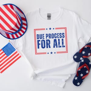 May include: White t-shirt with a blue and red graphic that says "Due Process For All". The graphic is surrounded by red and blue stars. The shirt is laid out on a white surface with a red, white, and blue hat and flip-flops.