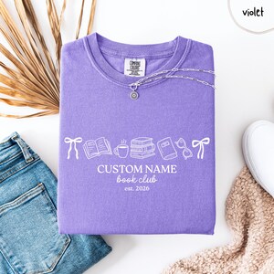 May include: A violet t-shirt featuring a white graphic print of bows, an open book, a coffee mug, a stack of books, a closed book, and glasses. The text reads CUSTOM NAME book club est. 2026. The shirt has a crew neckline and a Comfort Colors label.