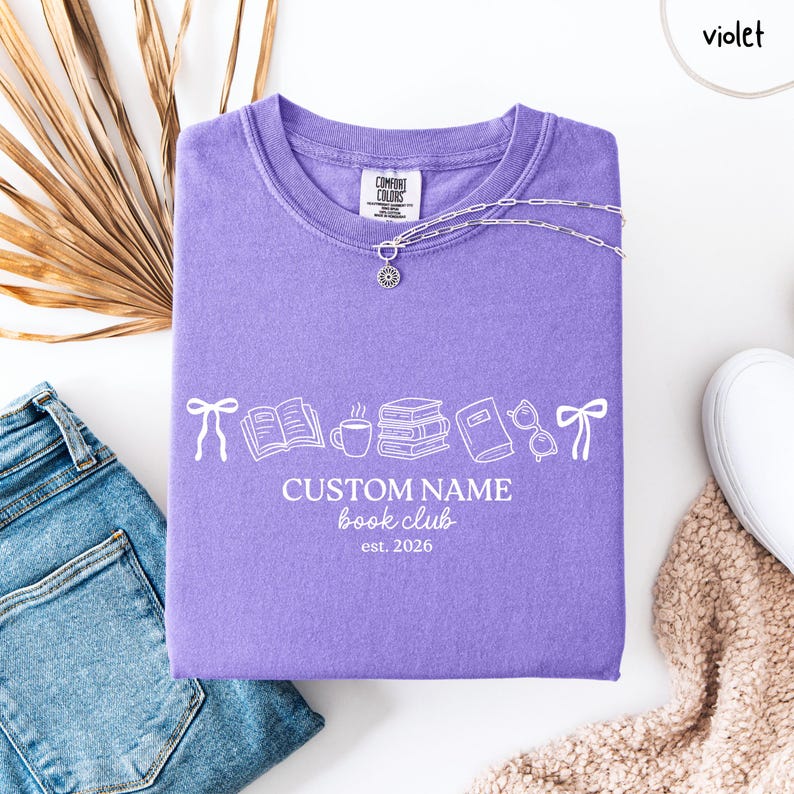 May include: A violet t-shirt featuring a white graphic print of bows, an open book, a coffee mug, a stack of books, a closed book, and glasses. The text reads CUSTOM NAME book club est. 2026. The shirt has a crew neckline and a Comfort Colors label.
