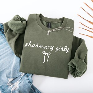 Puede incluir: Una sudadera verde oliva con las palabras "pharmacy girly" en escritura blanca y un lazo. Un collar plateado con un colgante est&aacute; sobre la sudadera. La sudadera se combina con unos vaqueros azules.