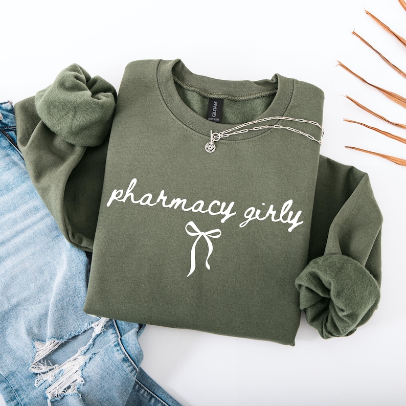 Puede incluir: Una sudadera verde oliva con las palabras "pharmacy girly" en escritura blanca y un lazo. Un collar plateado con un colgante est&aacute; sobre la sudadera. La sudadera se combina con unos vaqueros azules.
