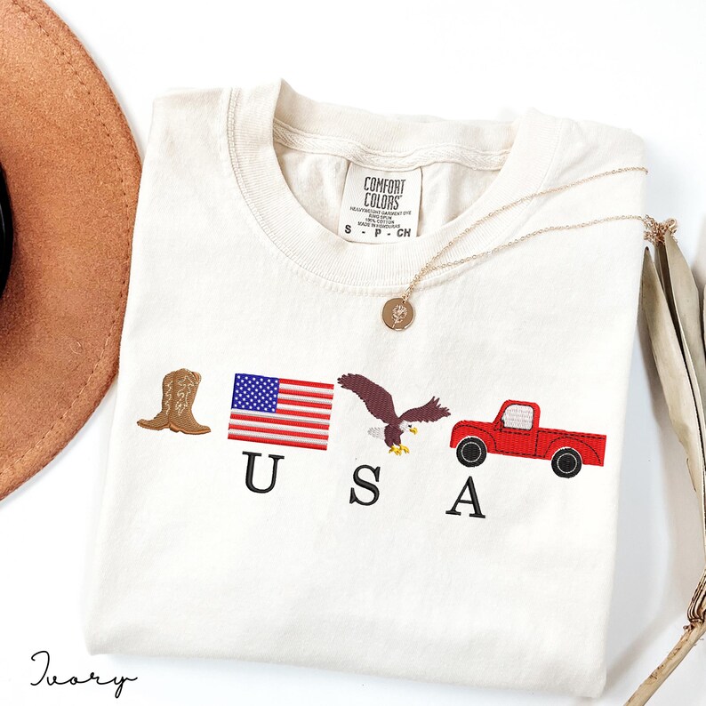 May include: Ivory-colored t-shirt with embroidered patriotic symbols: a cowboy boot, American flag, eagle, and red truck. The word "USA" is embroidered below the symbols. A gold necklace and a brown hat are also visible.