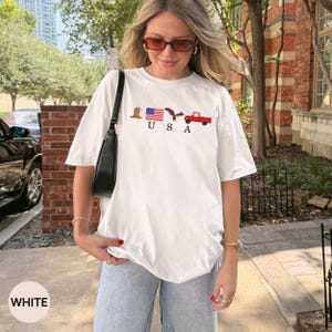 May include: White t-shirt with a patriotic USA design. The graphic includes a cowboy hat, American flag, eagle, and a red pickup truck. The word "USA" is printed below the symbols.