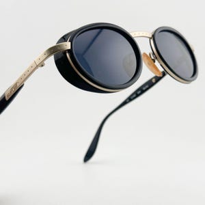 May include: Black and gold round sunglasses with a unique design. The sunglasses have a black frame with a gold metal band around the lenses. The arms of the sunglasses are black with gold accents.