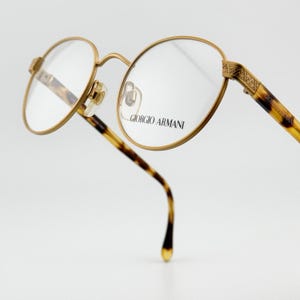 Puede incluir: Gafas con montura dorada y lentes ovaladas. Las monturas presentan un patrón de carey en las patillas y el texto "GIORGIO ARMANI" está impreso en la lente. Sobre un fondo blanco.