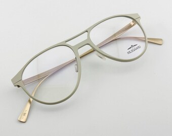 May include: A pair of eyeglasses with a light gray frame and gold-colored temples. The lenses are clear, and the brand name "KILSGAARD" is visible on the right lens. The glasses have a modern, minimalist design.