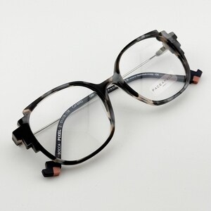 May include: Eyeglasses with a unique geometric design. The frames are a mix of black, gray, and tan, with clear lenses. The temples have a pixelated design, and the brand name "FACE A FACE" is visible on the lens.