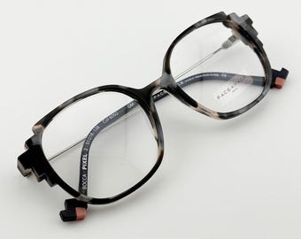 May include: Eyeglasses with a unique geometric design. The frames are a mix of black, gray, and tan, with clear lenses. The temples have a pixelated design, and the brand name "FACE A FACE" is visible on the lens.