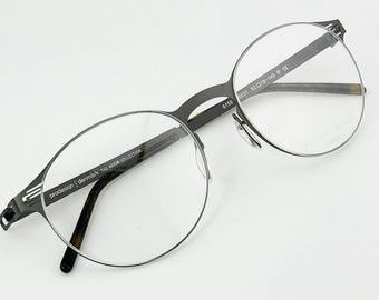 May include: A pair of eyeglasses with a dark gray metal frame and clear lenses. The glasses have a round shape and the text "prodesign: denmark THE AXIOM COLLECTION" is visible on the arm. The lenses are clear and the arms have a brown pattern.