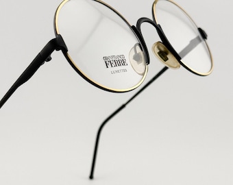 May include: Close-up of eyeglasses with round lenses. The frames are black with gold-colored rims. The brand name "GIANFRANCO FERRE LUNETTES" is printed on the lens. The glasses have a classic, elegant design.