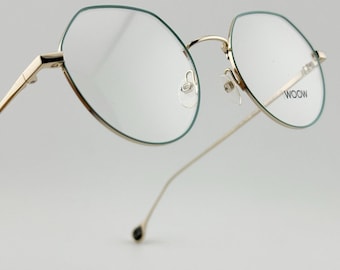 May include: Gold-toned eyeglasses with a thin frame and a unique geometric shape. The lenses are clear, and the rims have a subtle green outline. The text "WOOW" is visible on one of the lenses. The arms of the glasses are slender and elegant.
