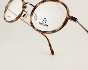 May include: Round eyeglasses with a brown tortoiseshell pattern on the frames. The metal arms and bridge are a light bronze color. The brand name "RODENSTOCK" is printed on the lens.