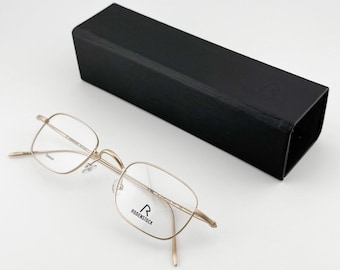 May include: Gold-tone eyeglasses with a rectangular frame and clear lenses, next to a black rectangular case. The glasses have the brand name "RODENSTOCK" printed on the lens.