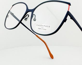 May include: A pair of eyeglasses with a dark blue frame and clear lenses. The frame has a unique, angular design with a red accent on the upper corner. The temple arms are a contrasting orange color. The text "FACE A FACE Paris Made in France" is visible.