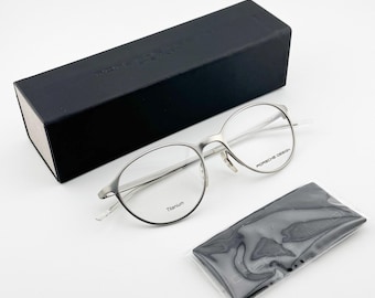 May include: Silver-toned titanium eyeglasses with round lenses, a black rectangular case, and a black cleaning cloth. The glasses have the words "Porsche Design" and "Titanium" printed on them.